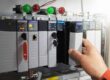 A close-up of an industrial control panel with multiple Programmable Logic Controllers (PLCs), where a technician’s hand is installing or removing a module.