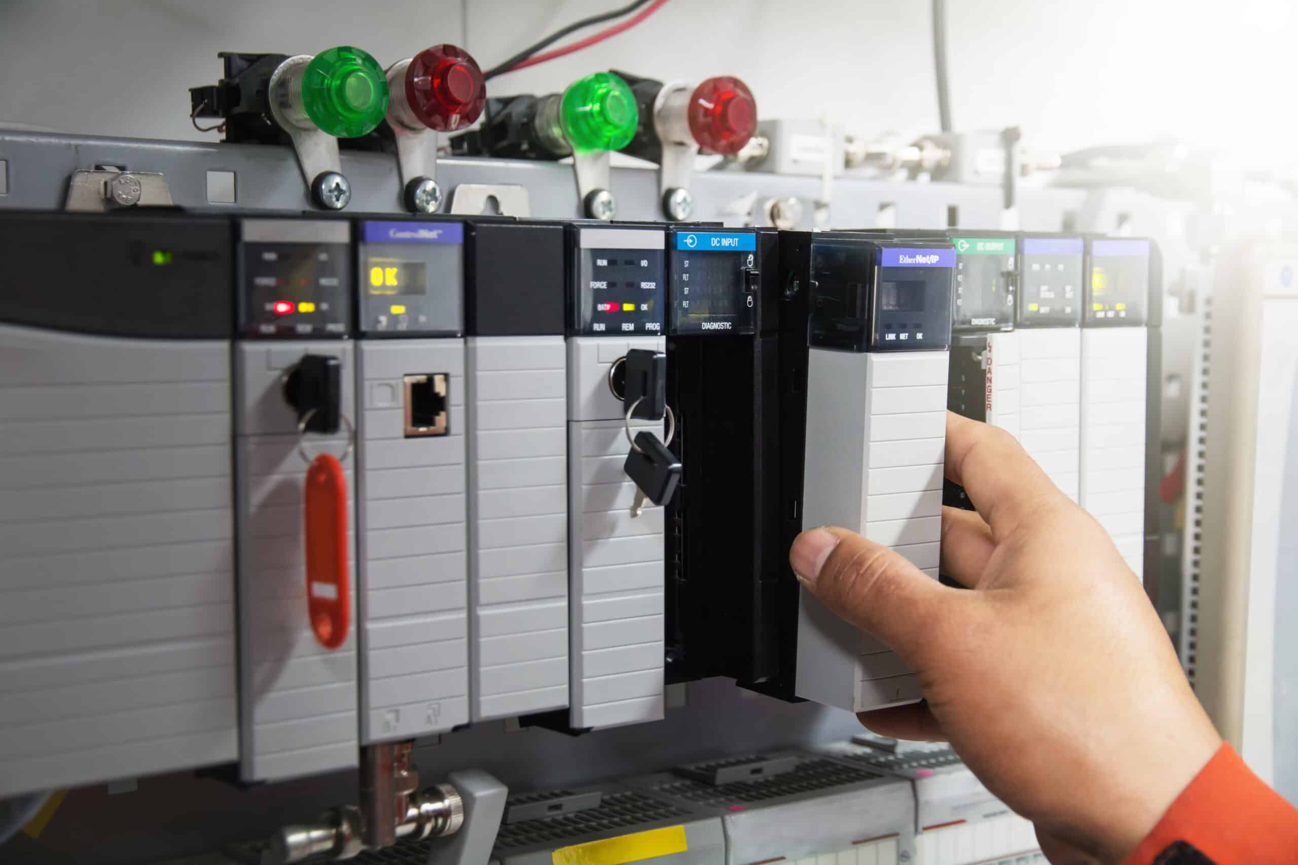 A close-up of an industrial control panel with multiple Programmable Logic Controllers (PLCs), where a technician’s hand is installing or removing a module.